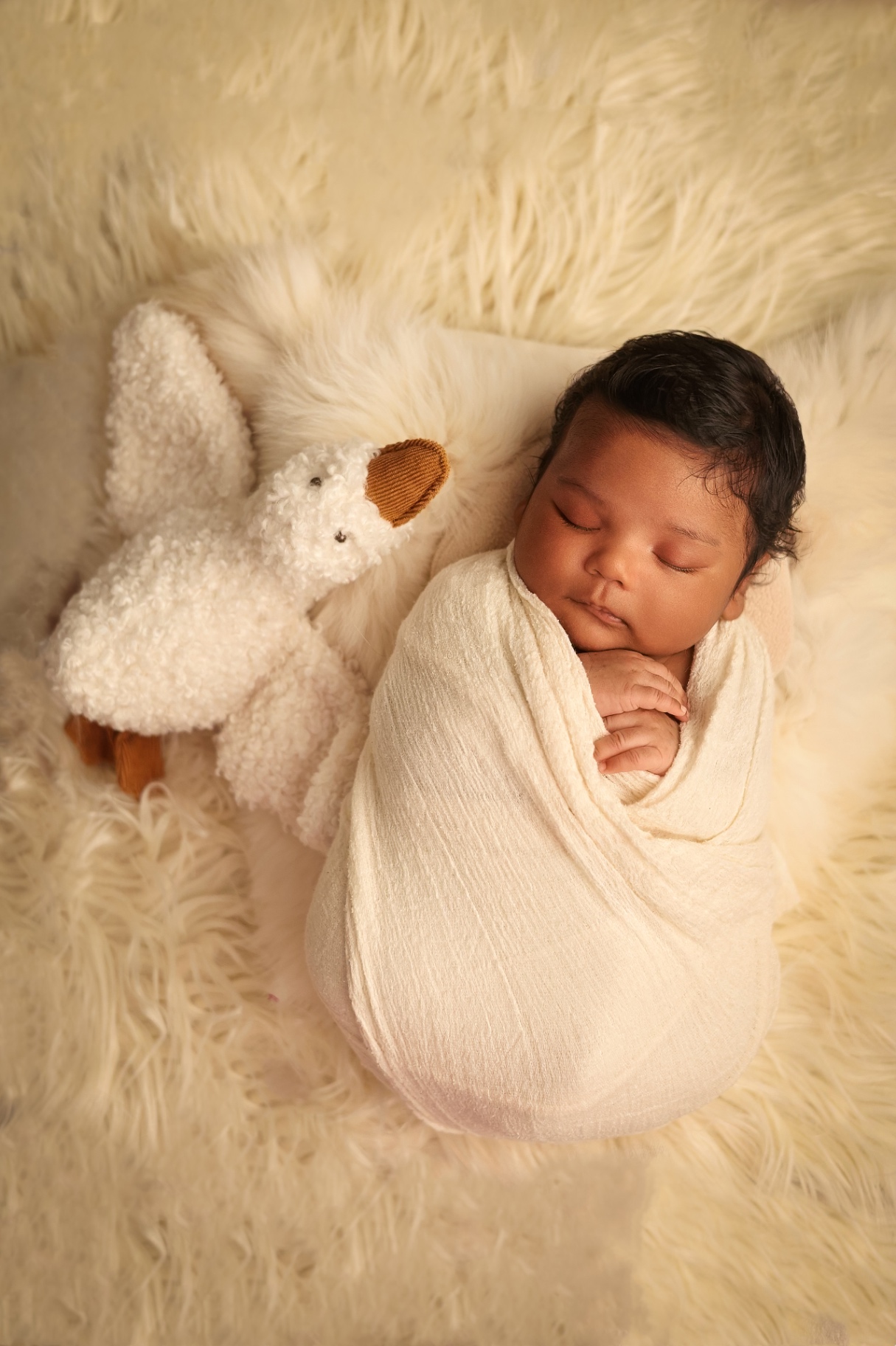 Newborn wrapped in cream on a soft fur rug beside a knitted bunny toy