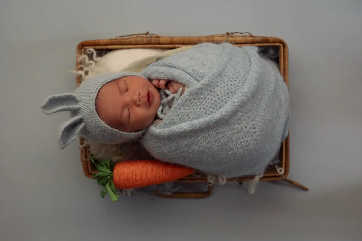 Sleeping newborn wrapped in blue knit with a bunny bonnet and a knitted carrot in a wicker basket