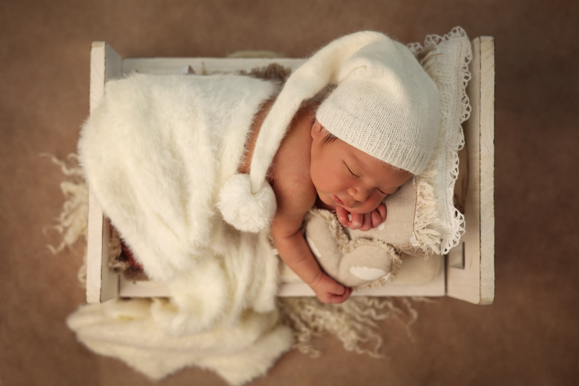 Newborn in a white sleepy cap resting in a tiny wooden bed with teddy