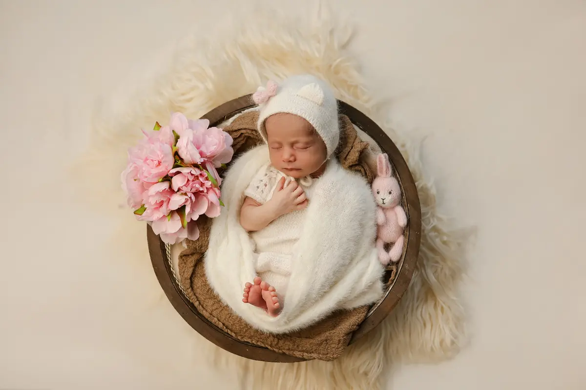 Newborn baby sleeping in a wooden bowl with pink peonies and a knit bunny