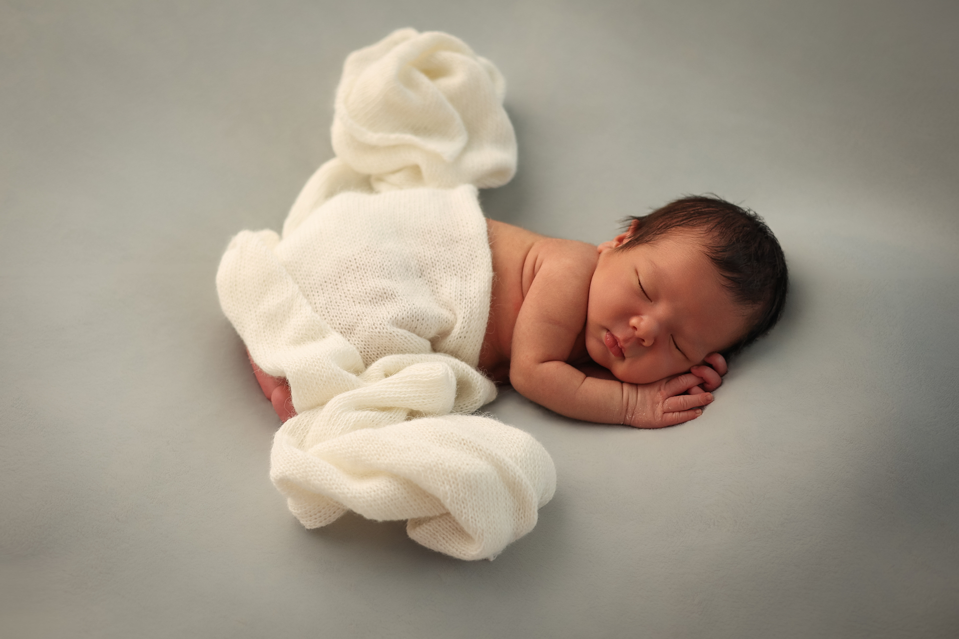 Sleeping newborn wrapped in a soft cream knit on a grey backdrop