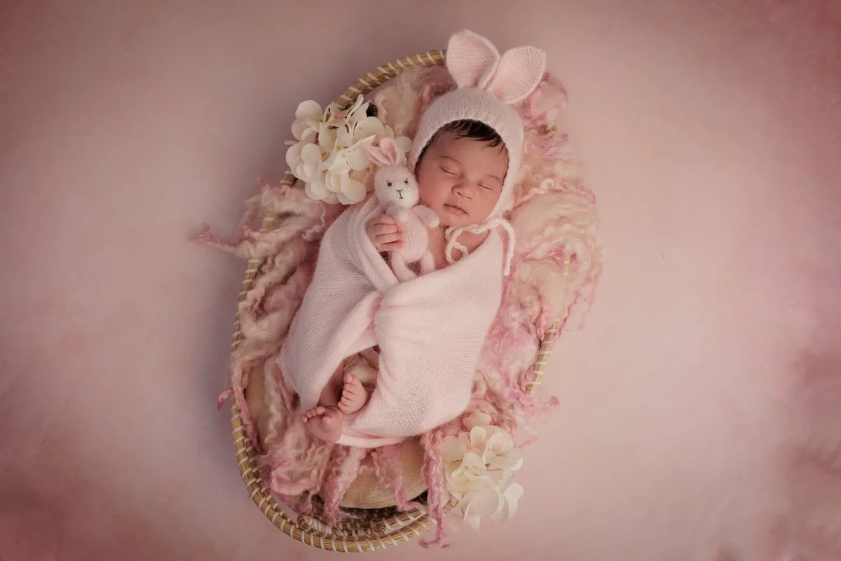 Newborn in a bunny-ear bonnet wrapped in pink, asleep in a basket of roses — taken in a Hendon nursery in soft morning light