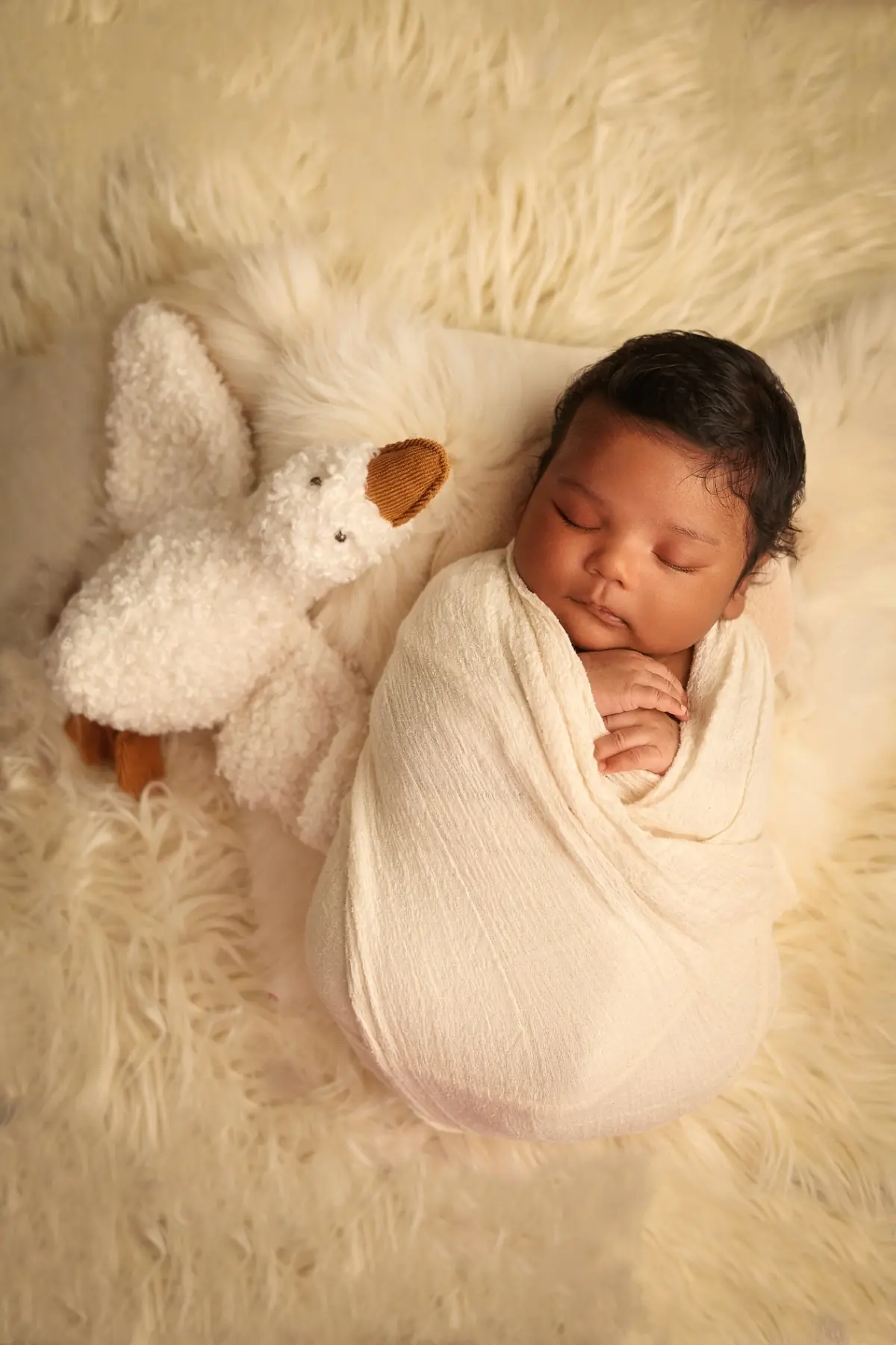 Newborn wrapped in cream on a soft fur rug beside a knitted bunny toy