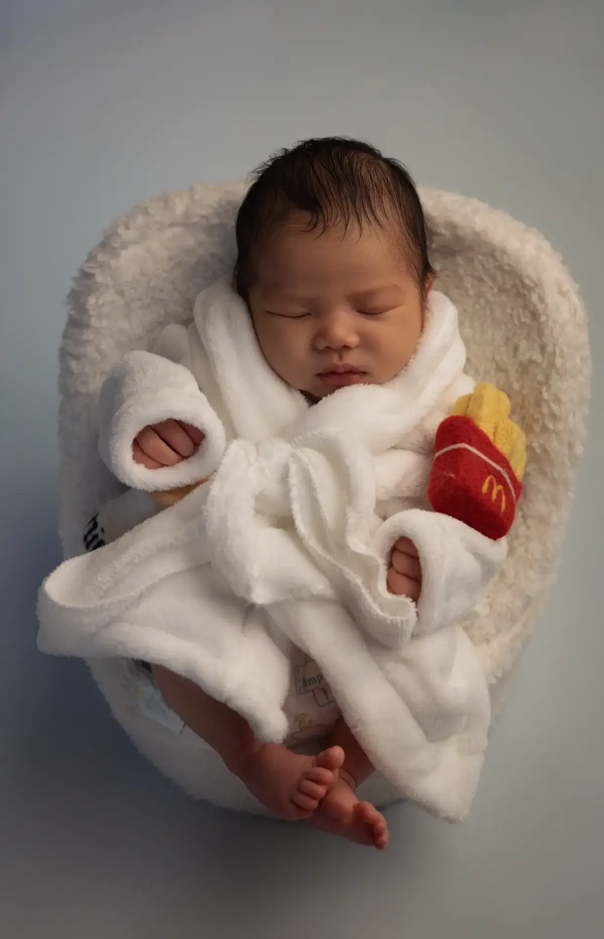Newborn sleeping in a tiny white bathrobe with a felt french-fries toy in a fluffy basket