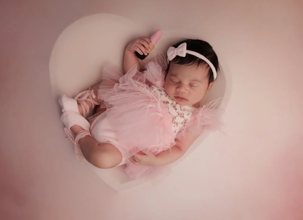 Newborn girl in a pink tutu and bow headband asleep on a soft white heart backdrop