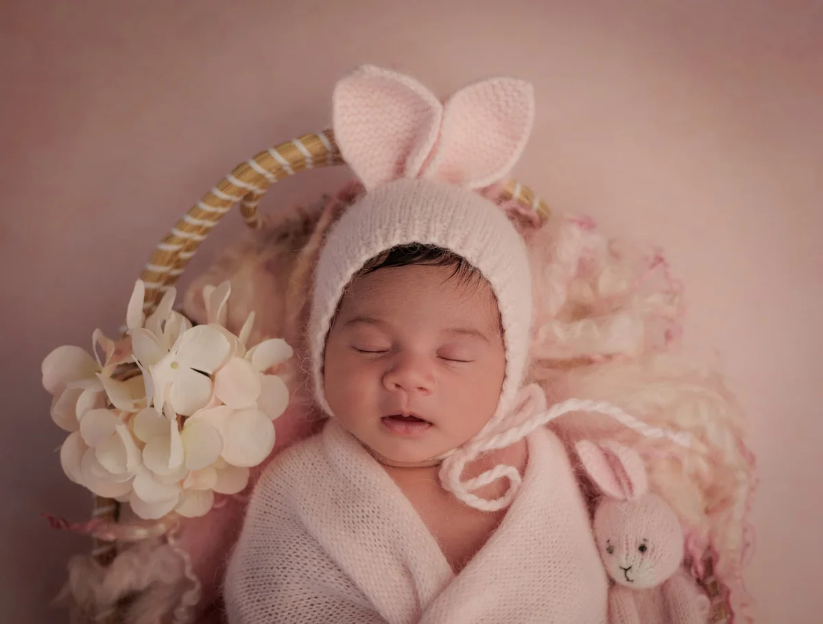 Newborn in a bunny-ear bonnet and cream wrap among pink flowers with a knitted bunny — photographed at home in West Hampstead NW6