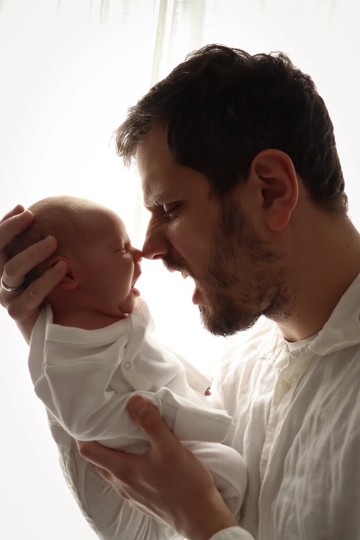 Father and yawning newborn touching noses in soft daylight