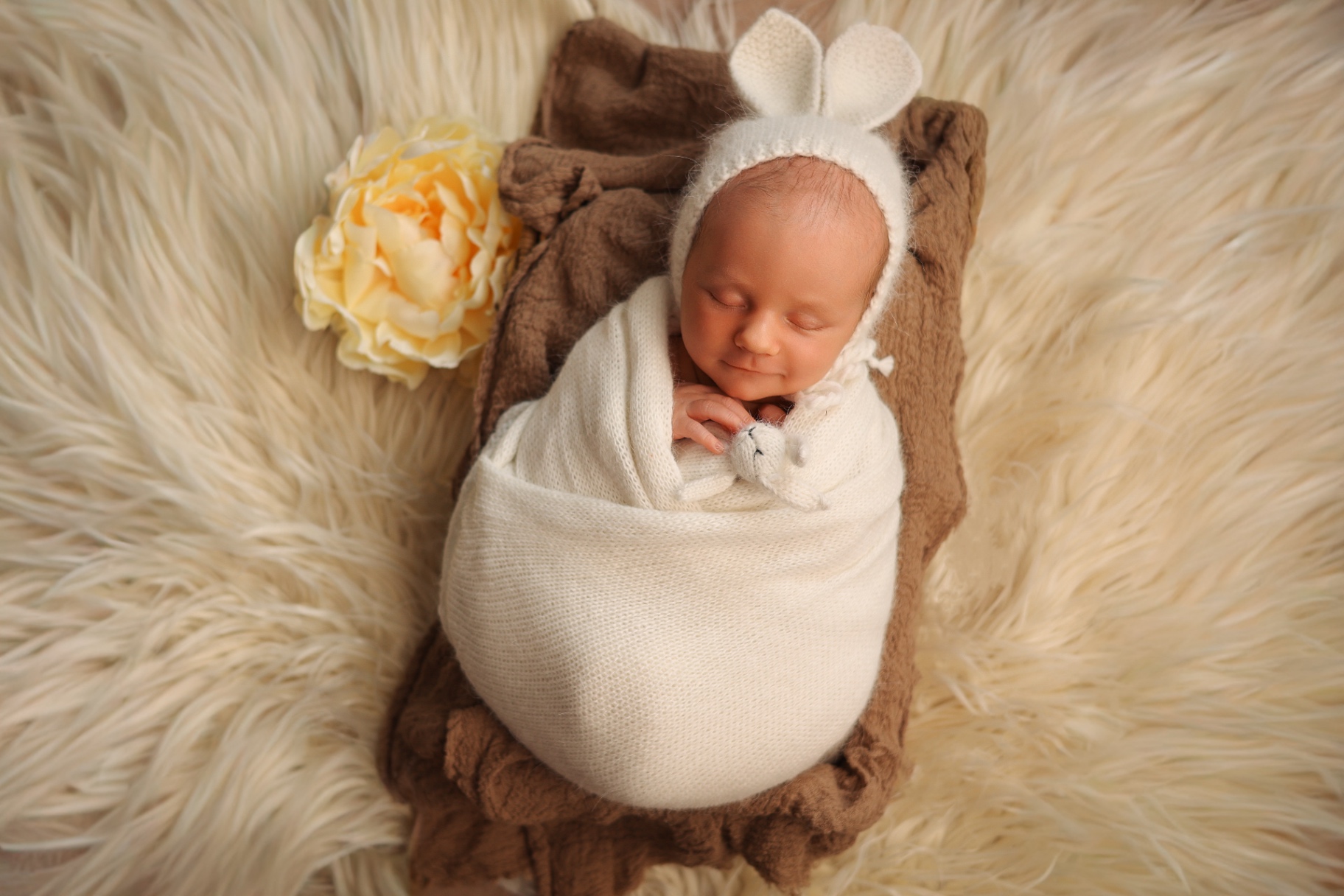 Smiling newborn in a white bunny-ear bonnet wrapped in cream beside a yellow peony