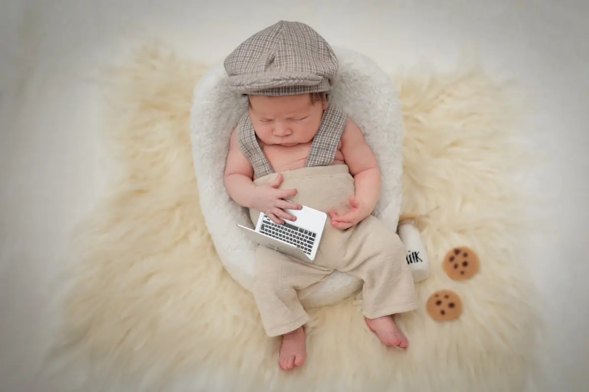 Newborn in a flat cap and braces resting with a tiny laptop prop