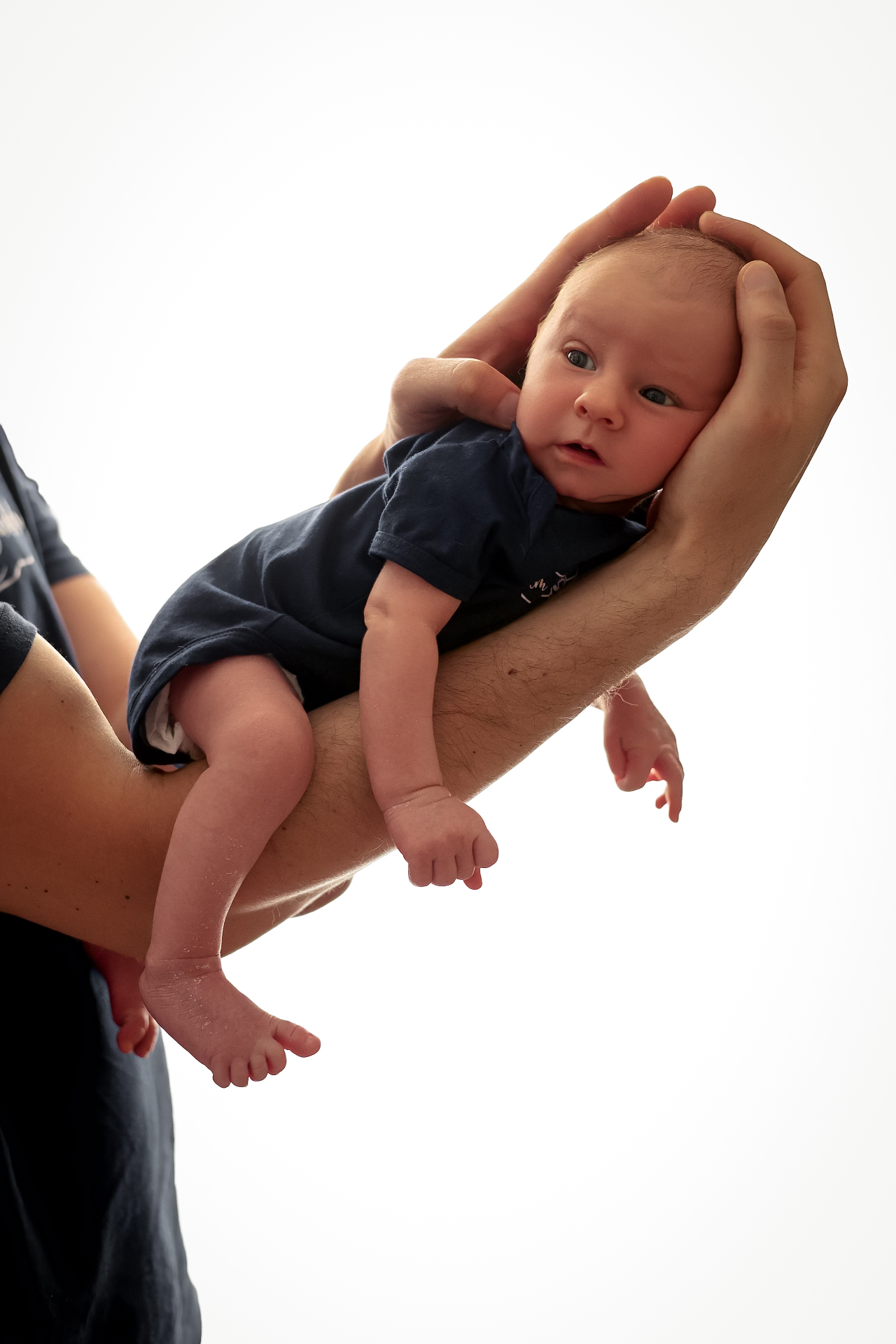 Father holding his awake newborn baby aloft against a bright white backdrop