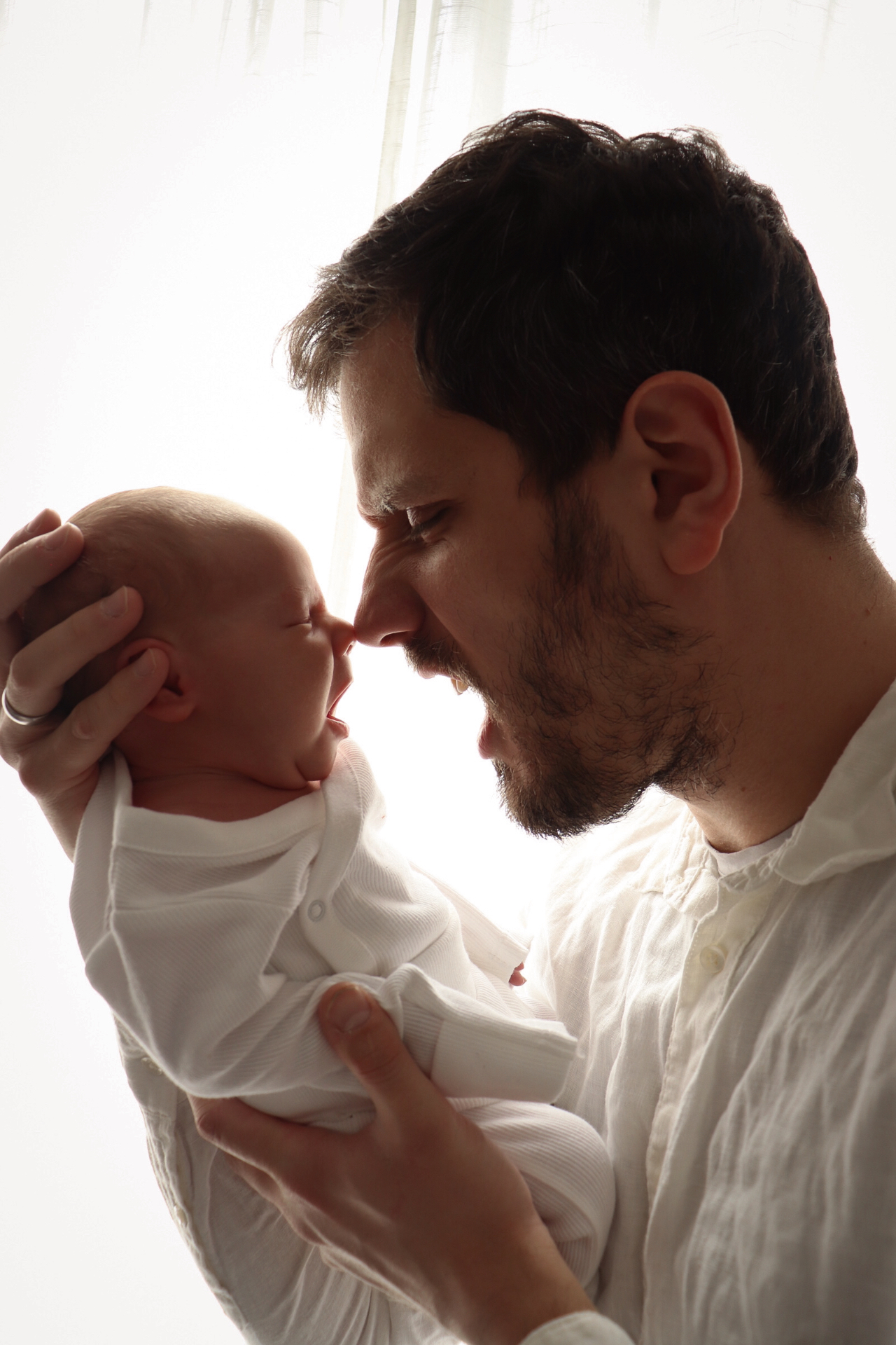 Father and yawning newborn touching noses in soft daylight