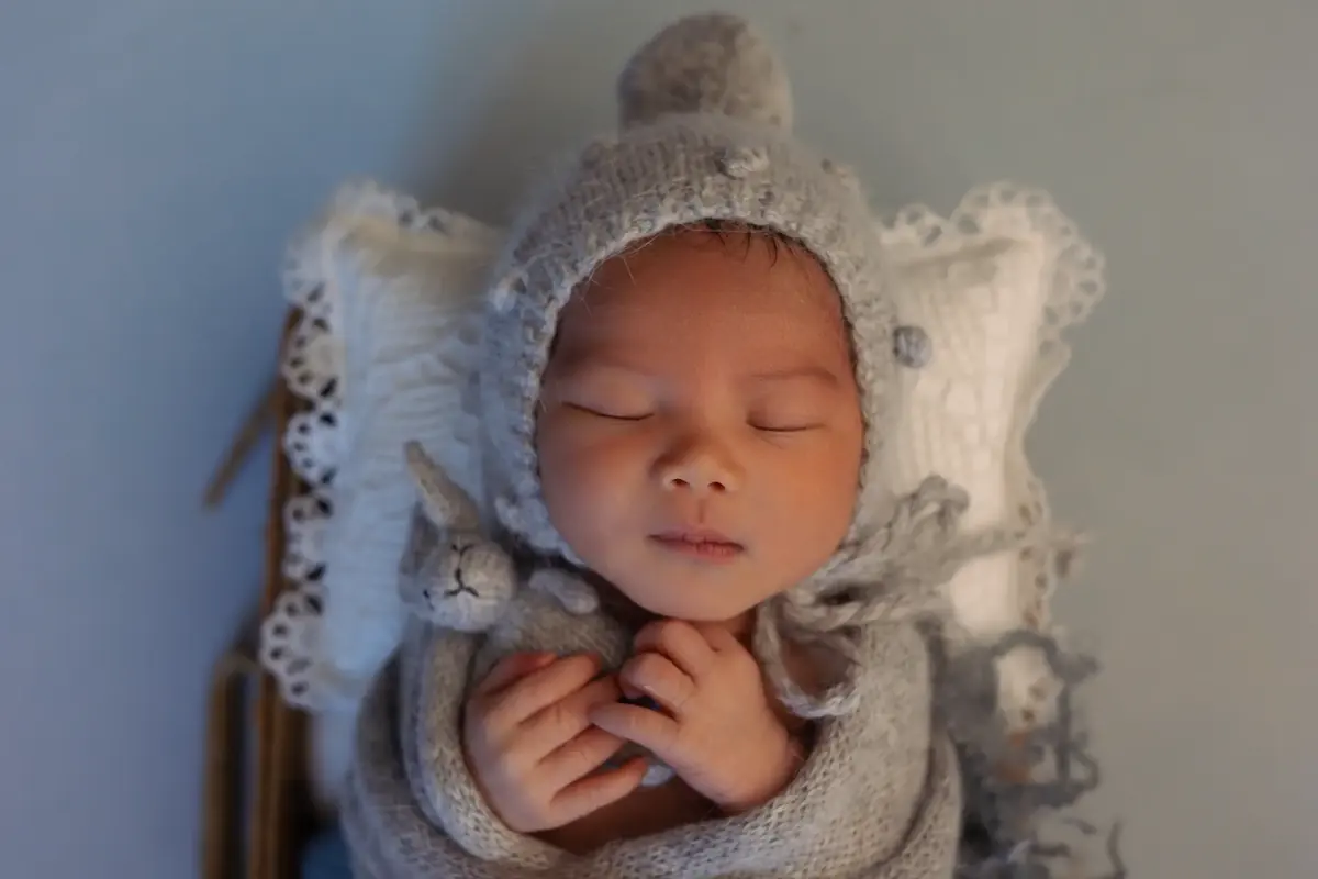 Sleeping newborn in a grey knit bonnet with a pom-pom cuddling a tiny knitted rabbit