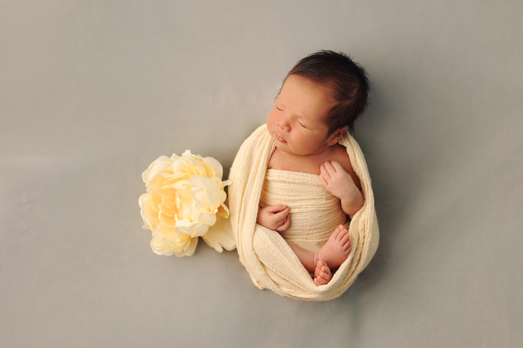 Newborn wrapped in cream fabric beside a soft yellow peony