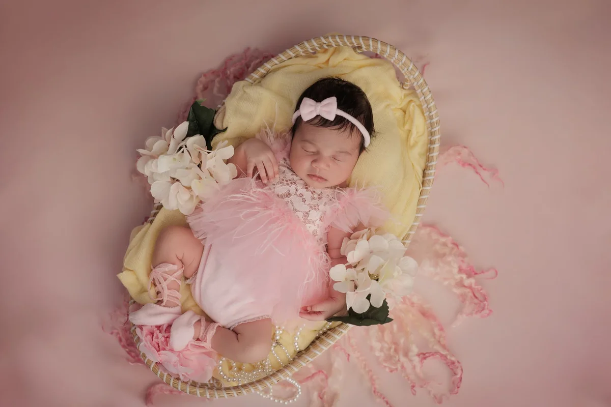 Newborn in a pink tutu asleep in a heart-shaped basket lined with yellow and roses — photographed with an older sibling at home in Colindale
